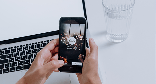A person holding a smartphone displaying the Canva app with the text 'Empowering the world to design,' placed near a laptop and a glass of water on a desk.