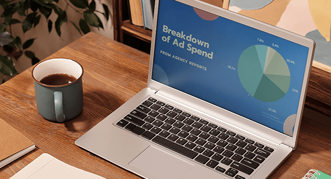 A laptop on a wooden desk displaying a report titled 'Breakdown of Ad Spend' with a pie chart, accompanied by a coffee mug, notebook, and plant.
